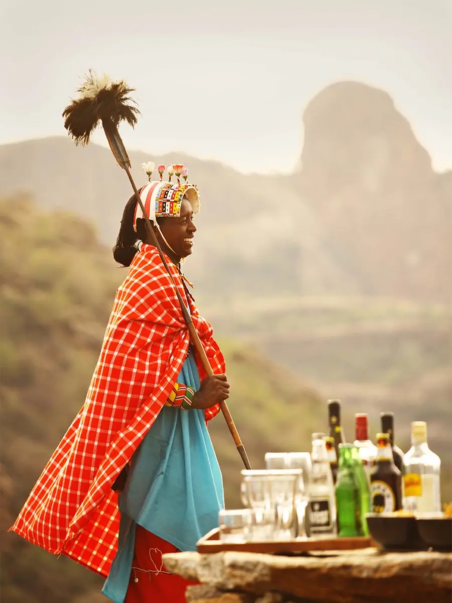 A Maasai host in a red shuka beside a rock-top bar above a hazy valley